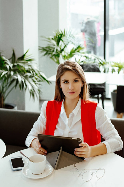 The strict business lady sits at a little table in cafe, uses the Internet on the tablet and has coffee. The woman in a white shirt and a red jacket during a lunch in business center. - Foto, Imagen