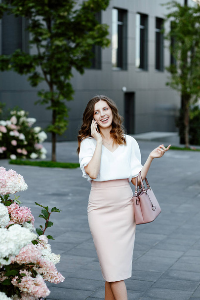 Office style. A portrait of the young positive business lady in the white blouse talking on the smartphone against the background of business center. - Fotó, kép