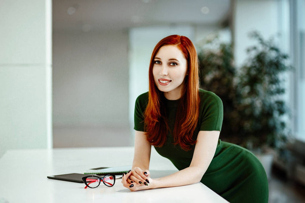 Business portrait of the beautiful red-haired girl in a green dress against the background of office. - Foto, Imagem