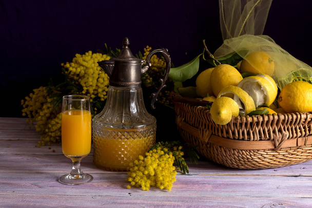 Stilleven met verse mimosa takken, kruik en glas met SAP en citroenen in een rieten mand op een houten tafel close-up - Foto, afbeelding