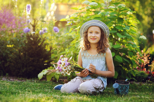 happy child girl playing little gardener in summer, wearing funny hat and holding bouquet of flowers. Sitting on green lawn, enjoying summertime vacations outdoor - Φωτογραφία, εικόνα