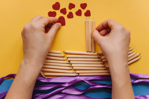 Το κορίτσι κάνει ένα δώρο με τα χέρια της με τη μορφή ενός πλοίου μολύβια και στέλνει ένα γράμμα με μια καρδιά. Δημιουργικό εργασιακό χώρο. Προετοιμασία για τις διακοπές. - Φωτογραφία, εικόνα