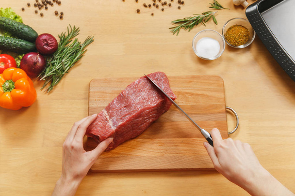 Raw meat. Fresh beef tenderloin on cutting board on wooden table with different vegetables, spices, mushrooms, baking tray, knife. Top view flat lay Copy space for advertisement. Woman cuts with knife - Photo, Image