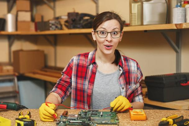 電気技師のマルチメータです。黄色の手袋で、かなり若い女性、メガネ デジタル電子エンジニアのさまざまなツールと木製のテーブルでのワーク ショップでの修復、はんだ付けコンピューター Pc のマザーボード. - 写真・画像