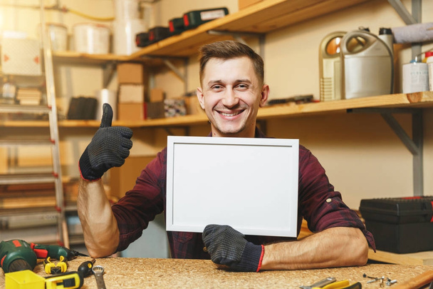 Handsome caucasian young man in plaid shirt, gloves working in carpentry workshop at wooden table place with blank frame, different tools. With empty place for text. Copy space for advertisement. - Photo, Image