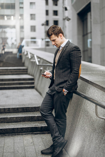 Carismático hombre de negocios elegante se encuentra al aire libre en el fondo de un centro de negocios con un teléfono en la mano y mira algo de información
 - Foto, Imagen