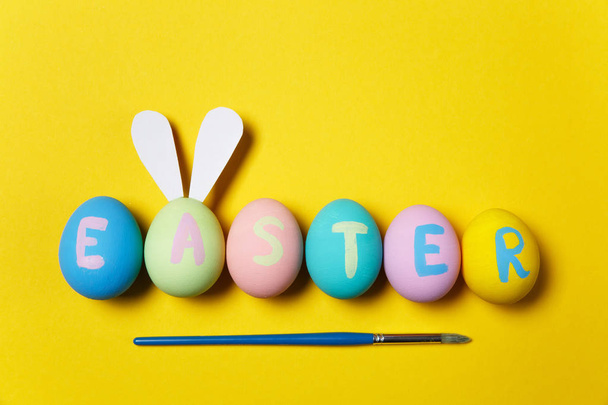 Row of colorful pastel monophonic painted Easter eggs with inscription Easter, fun bunny ears on egg, painting brush on yellow background. Copy space for advertisement. With place for text. Top view. - Foto, immagini