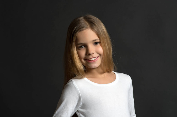 Beauty, look, hairstyle. Happy child, childhood concept. Girl with smile on adorable face on dark background. Youth, skincare, health. Kid model smiling with long healthy hair, copy space - Foto, imagen