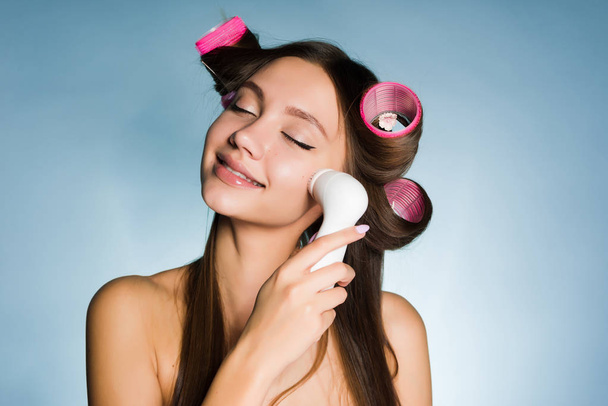 Uma mulher com rolos na cabeça limpa o rosto com uma escova para limpeza profunda
 - Foto, Imagem