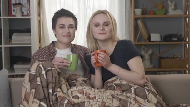 Duas jovens lésbicas estão sentadas no sofá, cobertas com um cobertor quente, segurando xícaras em suas mãos, bebendo chá escuro, café, abraçando 60 fps
 - Filmagem, Vídeo