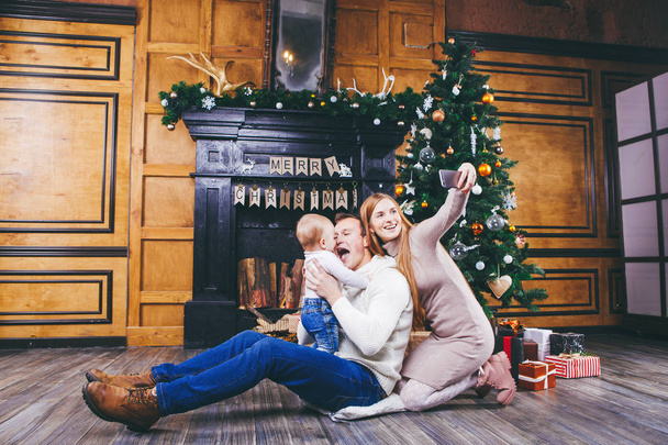 Christmas theme. young family with blond boy of one year sits on wooden floor against background of a Christmas tree with gifts and makes selfie, self-portrait on the front camera of a silver phone - Photo, Image