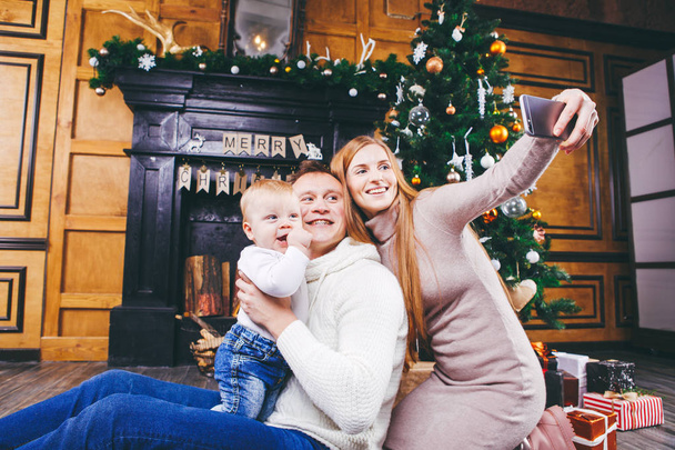 Christmas theme. young family with blond boy of one year sits on wooden floor against background of a Christmas tree with gifts and makes selfie, self-portrait on the front camera of a silver phone - Фото, зображення