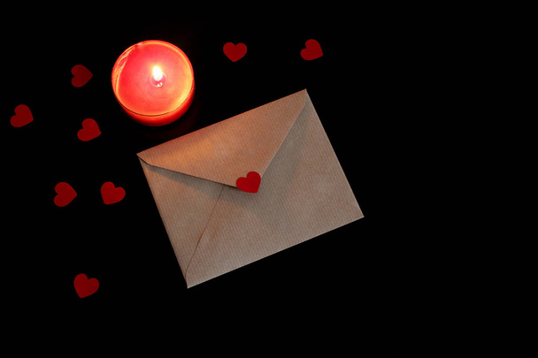 Valentines day or wedding mock-up scene with open craft paper envelope, paper hearts confetti and burning red candle on black background. Love concept. Flat lay, top view. Empty space for your text. - 写真・画像