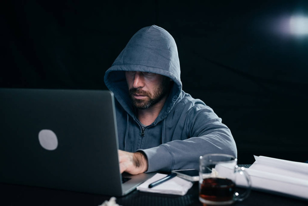 a man hacker in a sweatshirt with a hood sits behind a laptop and drinks tea - Foto, imagen