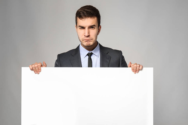 Emotional businessman with blank advertising board on grey background - Foto, Imagem