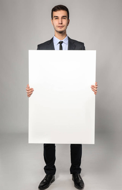 Handsome businessman with blank advertising board on grey background - Zdjęcie, obraz