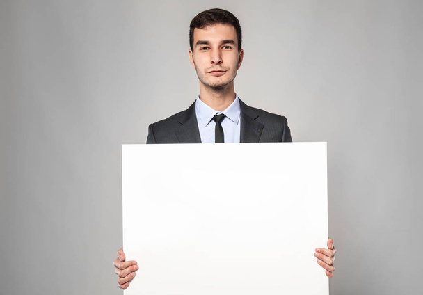 Handsome businessman with blank advertising board on grey background - Fotó, kép