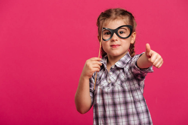 Cheerful little girl child holding fake glasses showing thumbs up. - 写真・画像