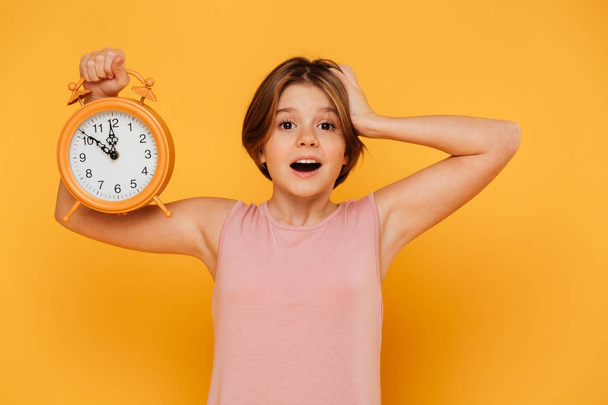 Surprised smiling girl showing alarm clock and holding her head isolated - Fotoğraf, Görsel