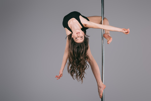 upside down of beautiful female dancer in sportswear exercising with pole and looking at camera on grey   - Photo, Image