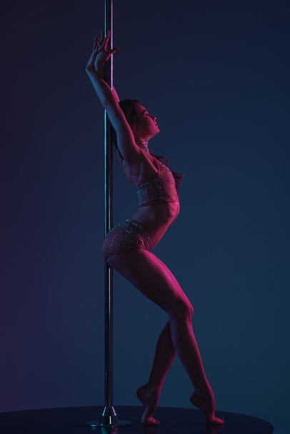 side view of seductive young female dancer leaning at pole on blue - Photo, Image