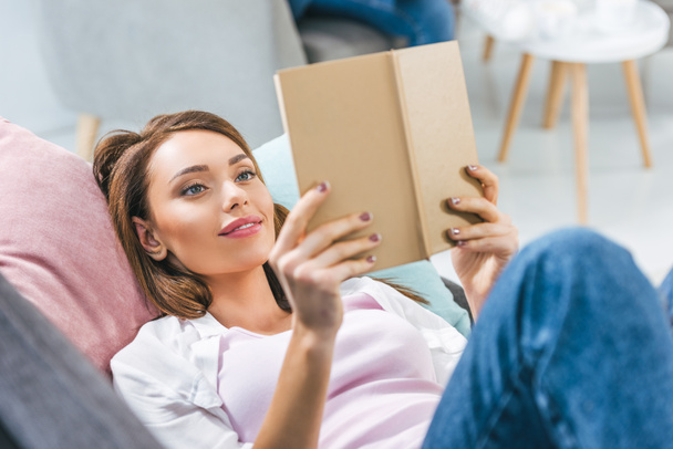 attractive girl reading book while lying on sofa at home - Fotografie, Obrázek