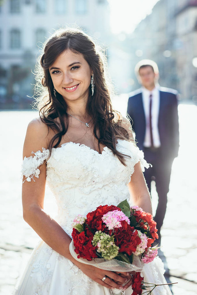 Smiling brunette bride with natural make-up is holding the wedding bouquet of red and pink flowers at the blurred background of the groom. Outdoor portrait. - Photo, Image
