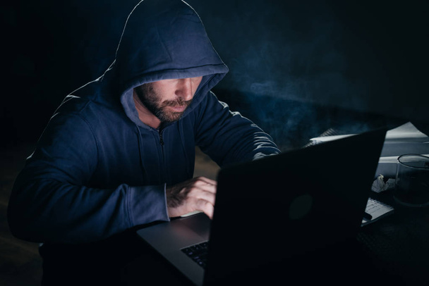 man hacker in a sweatshirt with a hood sits in the room behind a laptop - Фото, изображение