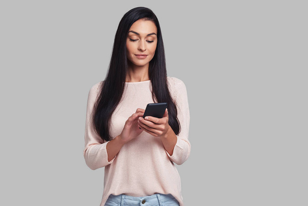 brunette woman using smart phone while standing against grey background - Φωτογραφία, εικόνα