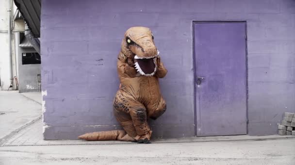 Homem em traje de dinossauro em pé inclina-se ao lado da porta na parede roxa
 - Filmagem, Vídeo