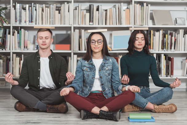 estudantes multiculturais meditando com livros sobre cabeças na biblioteca
 - Foto, Imagem