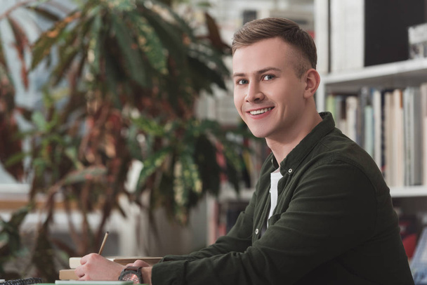 smiling male student sitting at table in library and looking at camera - Foto, Bild
