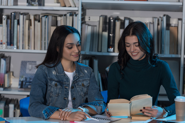 two multicultural female friends looking at book in library - Φωτογραφία, εικόνα