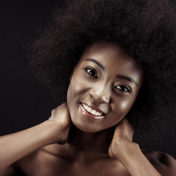 mujer afroamericana feliz sosteniendo su cuello y mirando a la cámara aislada en negro
 - Foto, Imagen