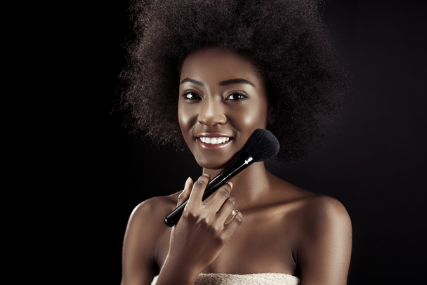 uśmiechający się african american kobieta robi makijaż z pędzla na czarnym tle - Zdjęcie, obraz