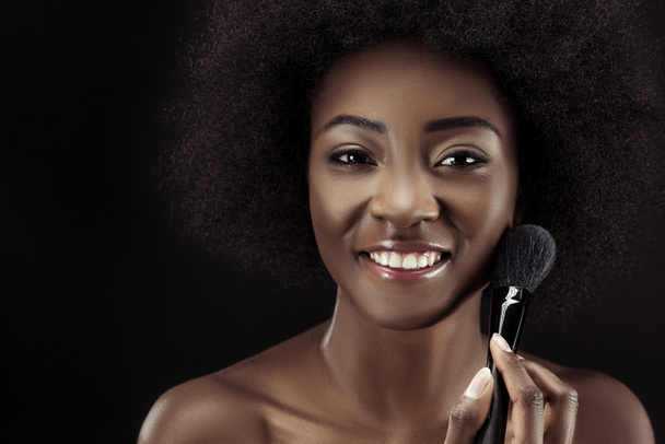 happy african american woman doing makeup with brush isolated on black - Fotografie, Obrázek