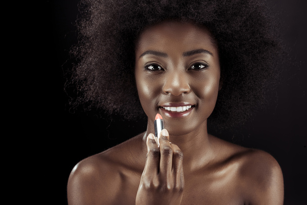 glückliche afrikanisch-amerikanische Frau, die Lippenstift aufträgt und isoliert auf schwarz in die Kamera schaut - Foto, Bild