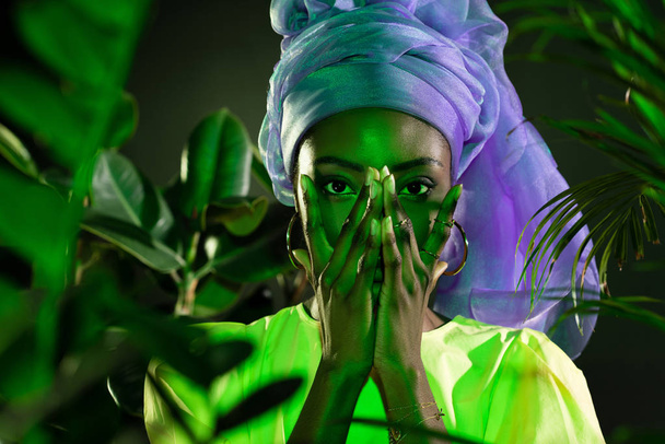 νεαρή αφρικανική αμερικανική γυναίκα στο παραδοσιακό σύρμα περιτύλιγμα κεφάλι που καλύπτουν το πρόσωπο με τα χέρια κάτω από το πράσινο φως - Φωτογραφία, εικόνα