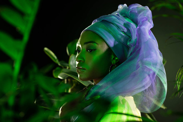 atractiva mujer afroamericana en envoltura de cabeza de alambre bajo luz verde detrás de hojas
 - Foto, imagen