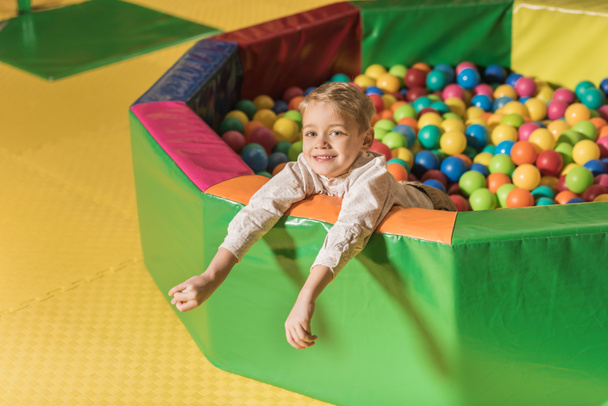 cute little boy smiling at camera while playing in pool with colorful balls  - Foto, Bild