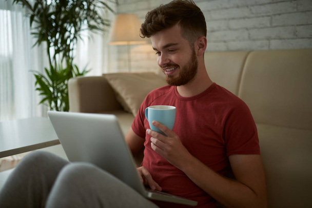 Mosolygó fiatal férfi csésze kávé néz video-ra laptop - Fotó, kép