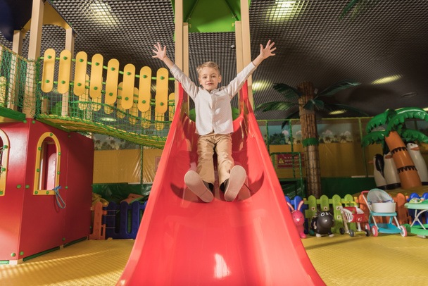 милий щасливий маленький хлопчик з піднятими руками грає на слайді в розважальному центрі
 - Фото, зображення