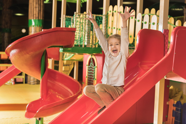 entzückender glücklicher kleiner Junge mit erhobenen Händen, der auf einer Rutsche im Spielzentrum spielt - Foto, Bild