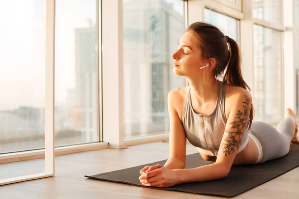 Portrait of young beautiful lady in sporty top and leggings lying on yoga mat and listening music in earphones while dreamily closing her eyes isolated - Photo, Image