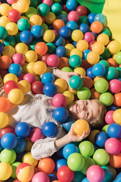 vista de ângulo alto do menino bonito sorrindo para a câmera enquanto deitado na piscina com bolas coloridas
 - Foto, Imagem