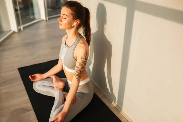 Retrato de dama en top deportivo y polainas sentadas en pose de loto y meditando sobre esterilla de yoga mientras escucha música en auriculares aislados en casa
 - Foto, imagen