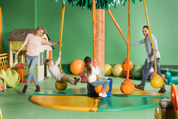 padres sonrientes mirando a niños felices balanceándose en columpios en el centro de entretenimiento
   - Foto, Imagen