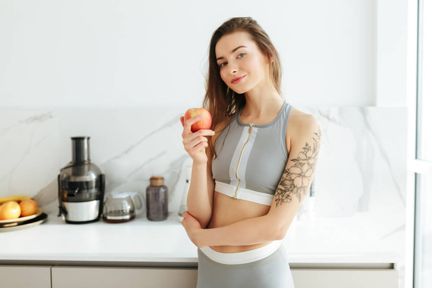 Retrato de bela senhora no topo desportivo em pé na cozinha e segurando maçã na mão enquanto sonhadoramente olhando na câmera isolada
 - Foto, Imagem
