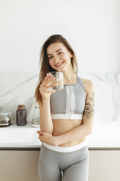 Portrait of beautiful lady in sporty top standing on kitchen and joyfully looking in camera with glass of water isolated - Foto, immagini