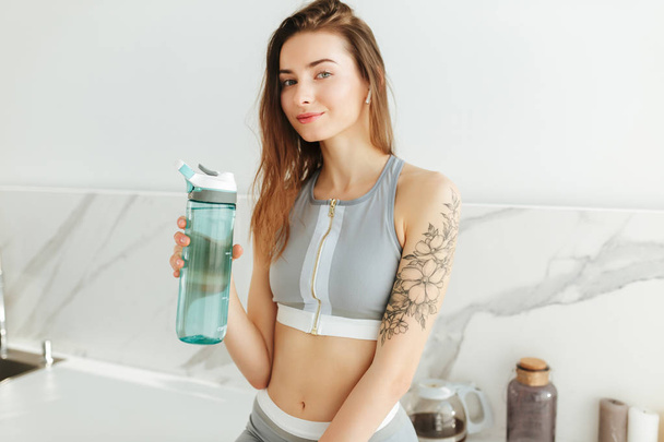 Portrait of beautiful girl in sporty top dreamily lookingin camera with bottle of water while standing on kitchen isolated - Фото, зображення